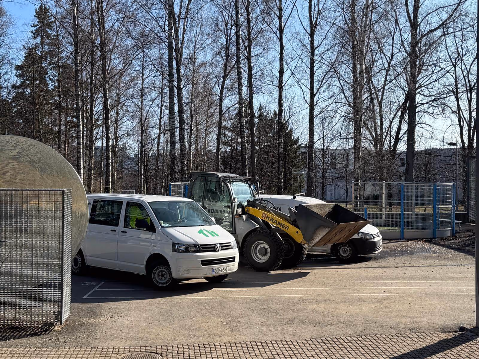 Ulkona on valkoinen pakettiauto, Kramer-pyöräkuormaaja ja toinen valkoinen auto pysäköitynä asfalttialueelle, taustalla puita ja rakennuksia aurinkoisessa säässä.