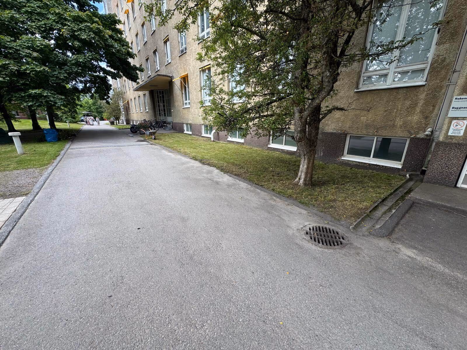 Ulkona on asfaltoitu tie, jonka vieressä on ruohoa ja rakennus. Rakennuksessa on ikkunoita ja puu. Valoisa sää.