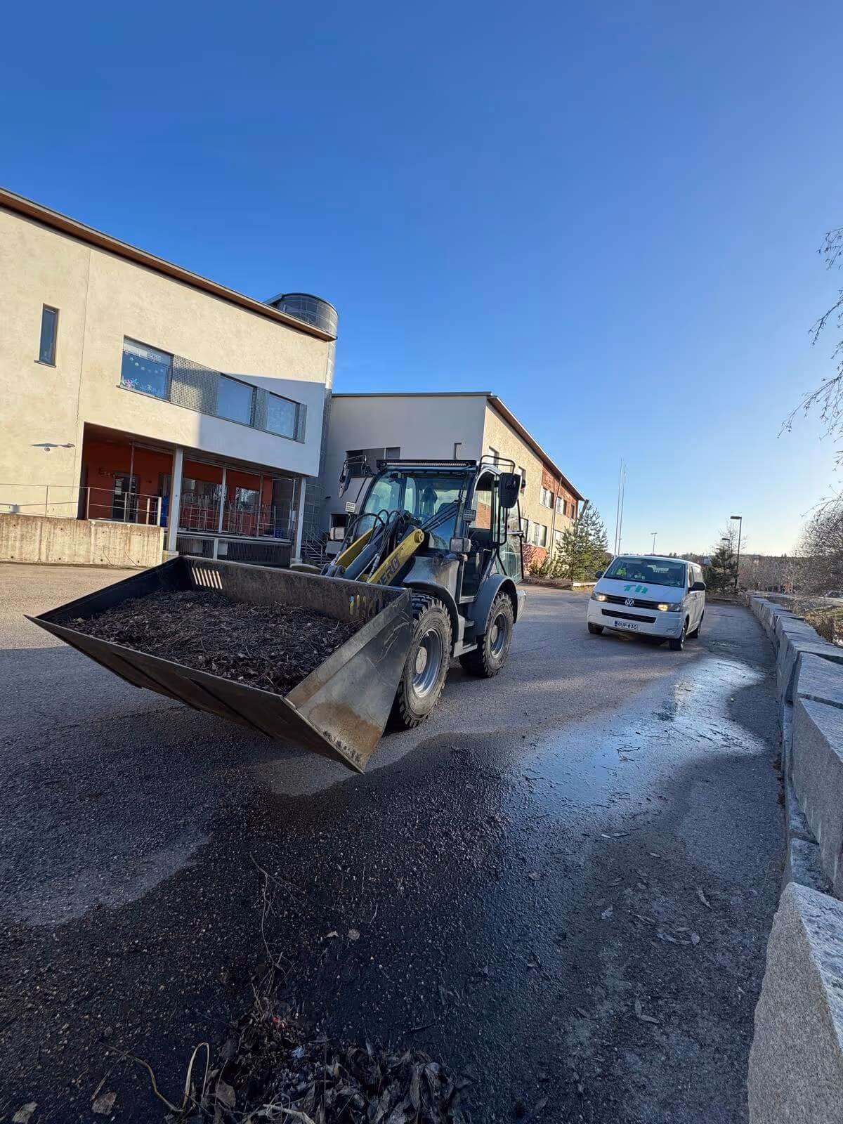 Etukuormaaja täynnä lehtiä ja oksia seisoo märällä asvaltilla rakennuksen edessä, valkoinen pakettiauto taustalla. Kirkas taivas.