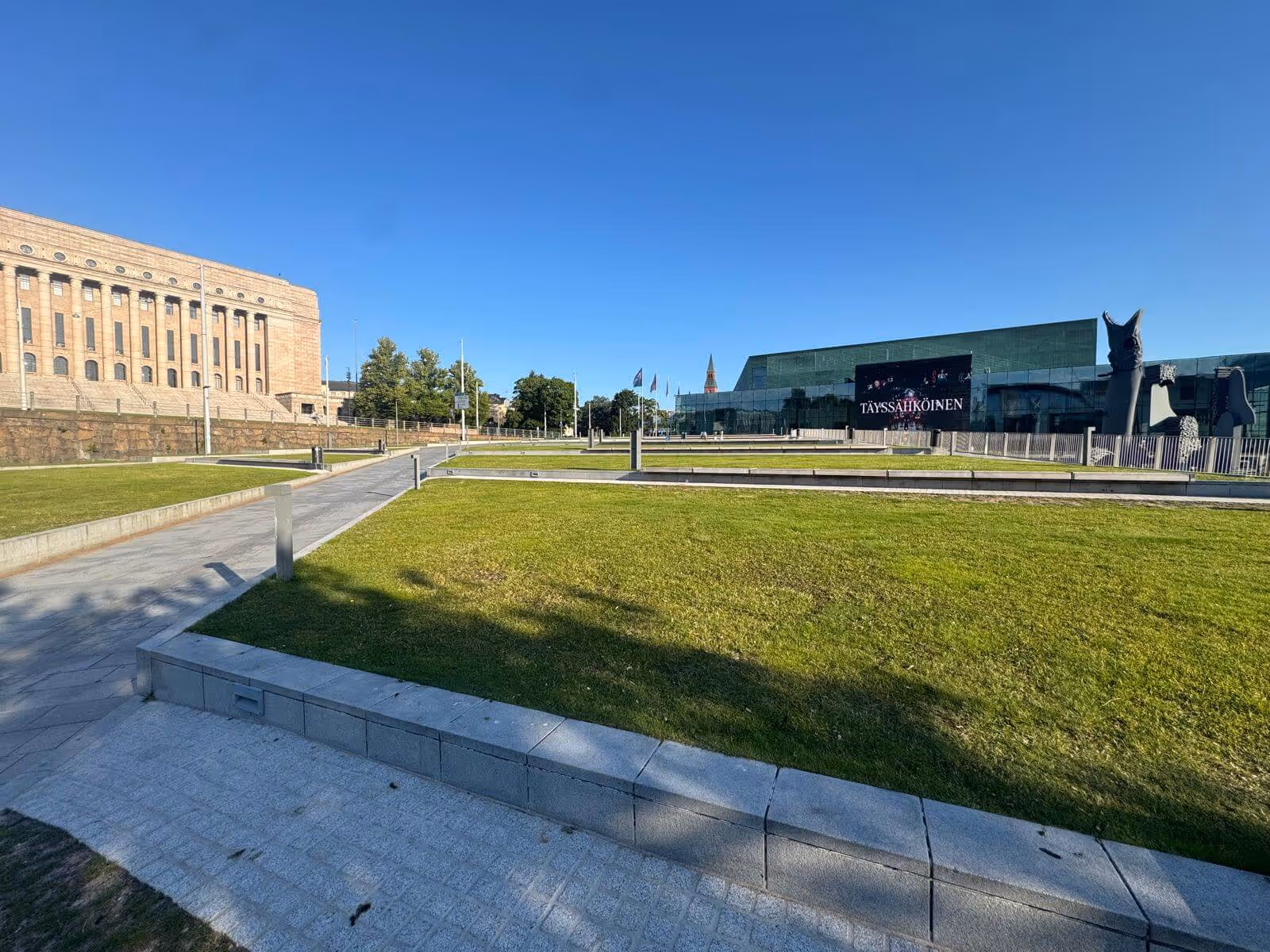 Näkymä Helsingin keskustassa: eduskuntatalo, Kiasma-museo, vihreä nurmikko ja sininen taivas.