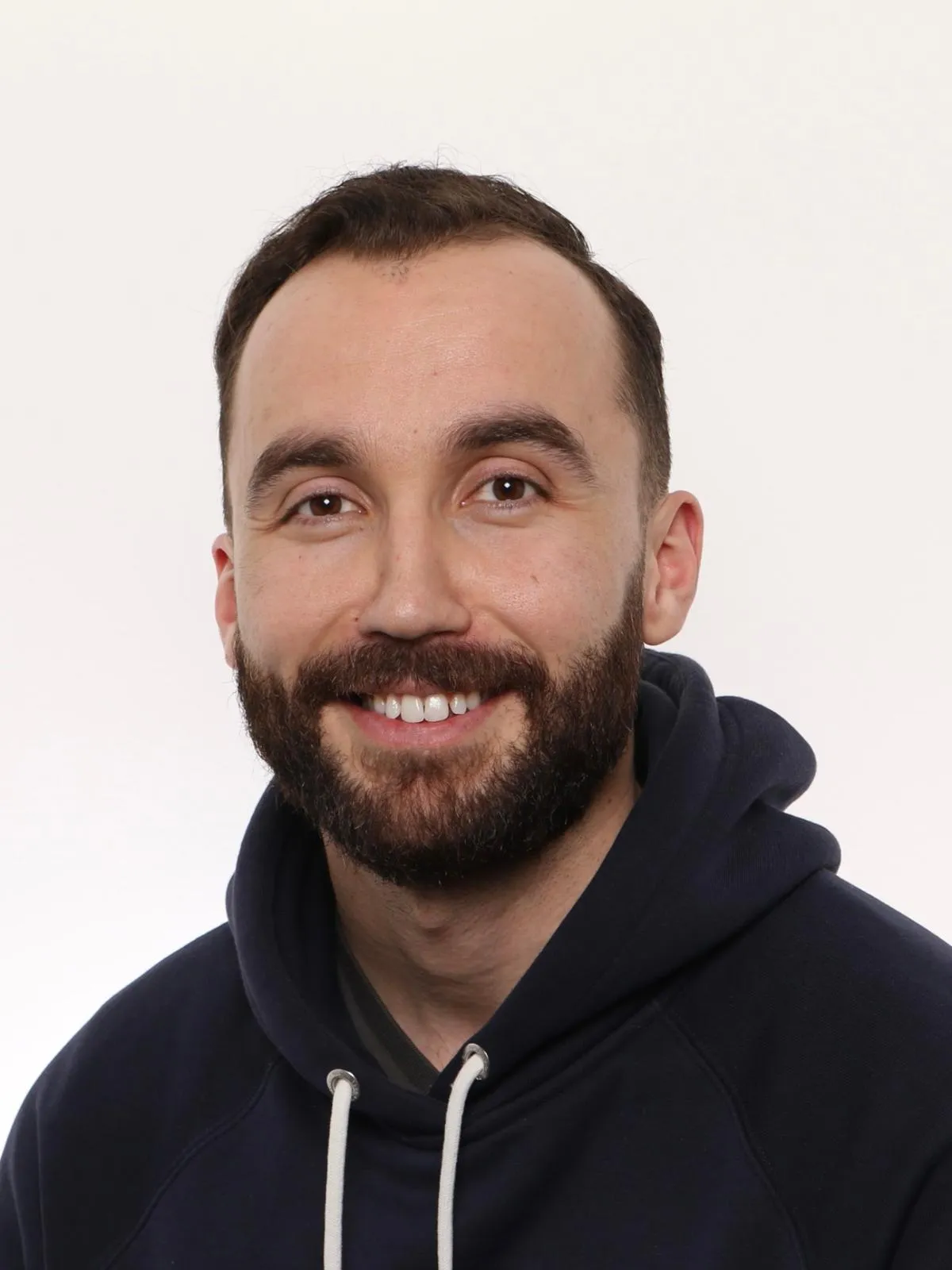 Lähikuva hymyilevästä miehestä tummansinisessä hupparissa, valkoisella taustalla.