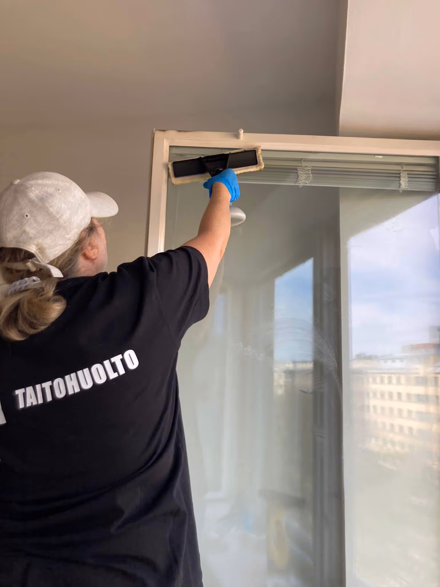 Nainen, jolla on musta paita, jossa lukee "TAITOHUOLTO", pesee ikkunaa sinisillä käsineillä ja ikkunanpesimellä.