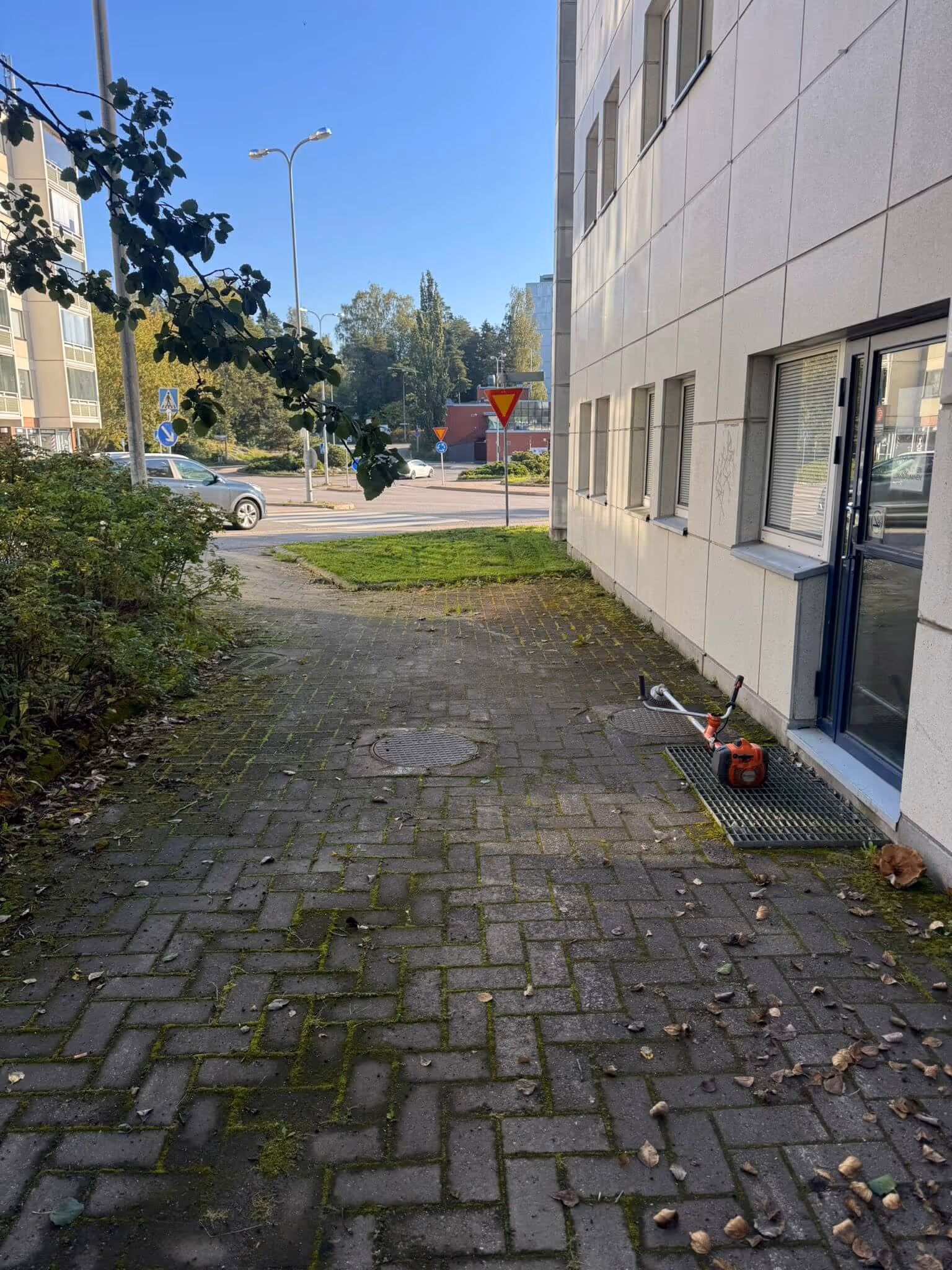 Ulkona on kivetty polku, jonka vieressä on rakennus ja jonka päällä on ruohotrimmeri. Polulla kasvaa sammalta ja siinä on lehtiä.