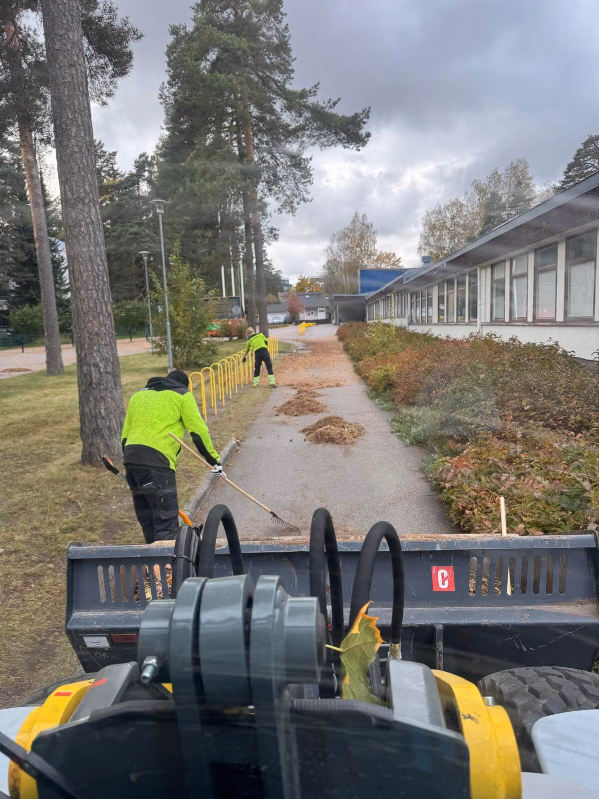 Kaksi henkilöä haravoi lehtiä polulta, toinen lähempänä kameraa ja toinen kauempana, molemmat käyttävät neonvihreitä takkeja, kuva on otettu ajoneuvosta, jossa näkyy musta kauha ja keltainen runko.