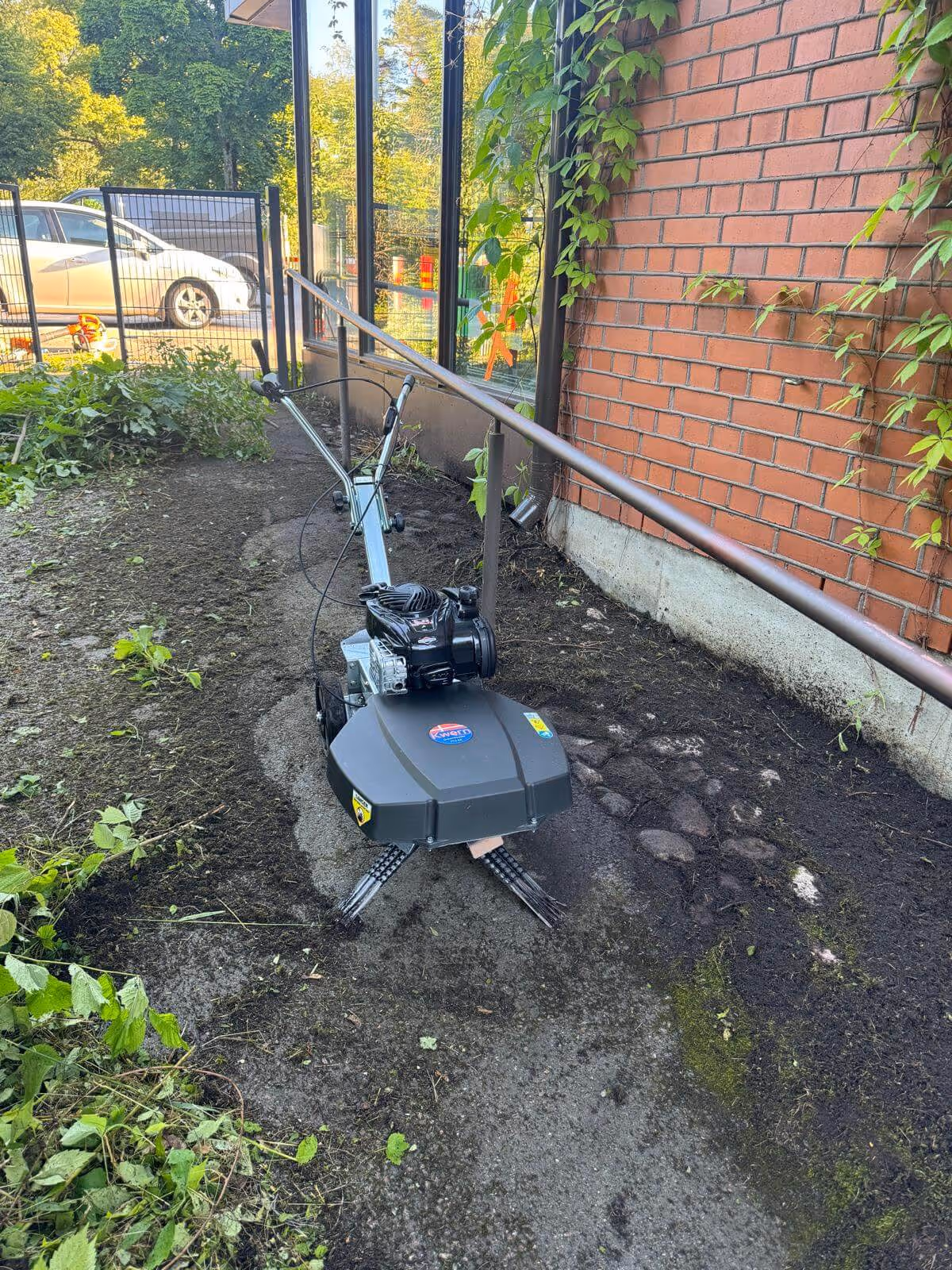 Maaperän muokkauskone seisoo maaperällä, jossa on vihreitä kasveja ja punatiilinen seinä taustalla.