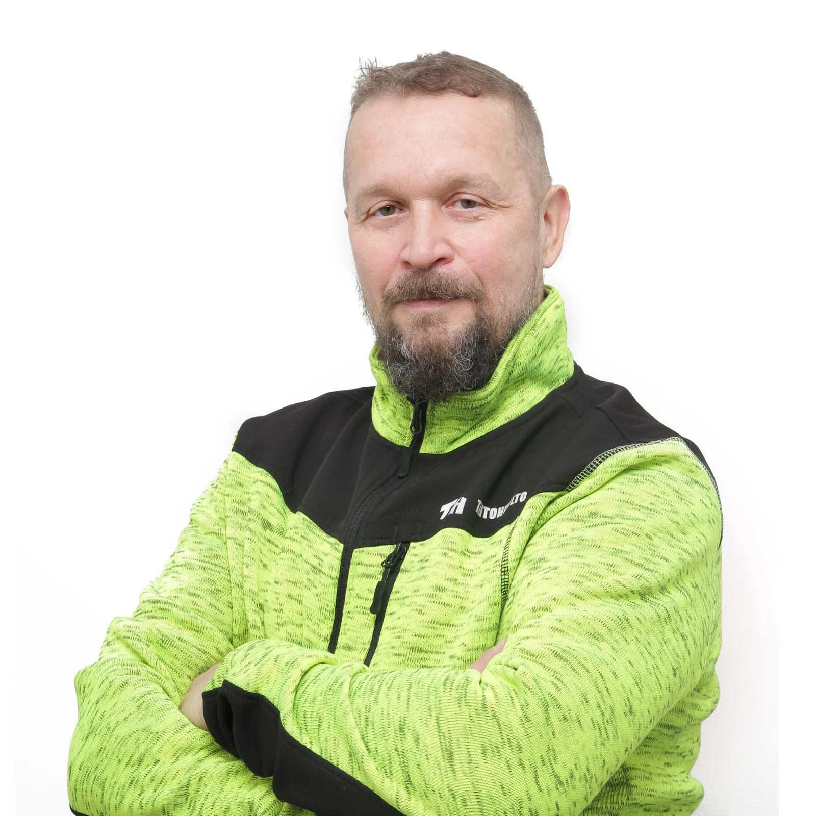 Keskellä oleva mies vihreässä ja mustassa takissa, kädet ristissä, valkoisella taustalla.