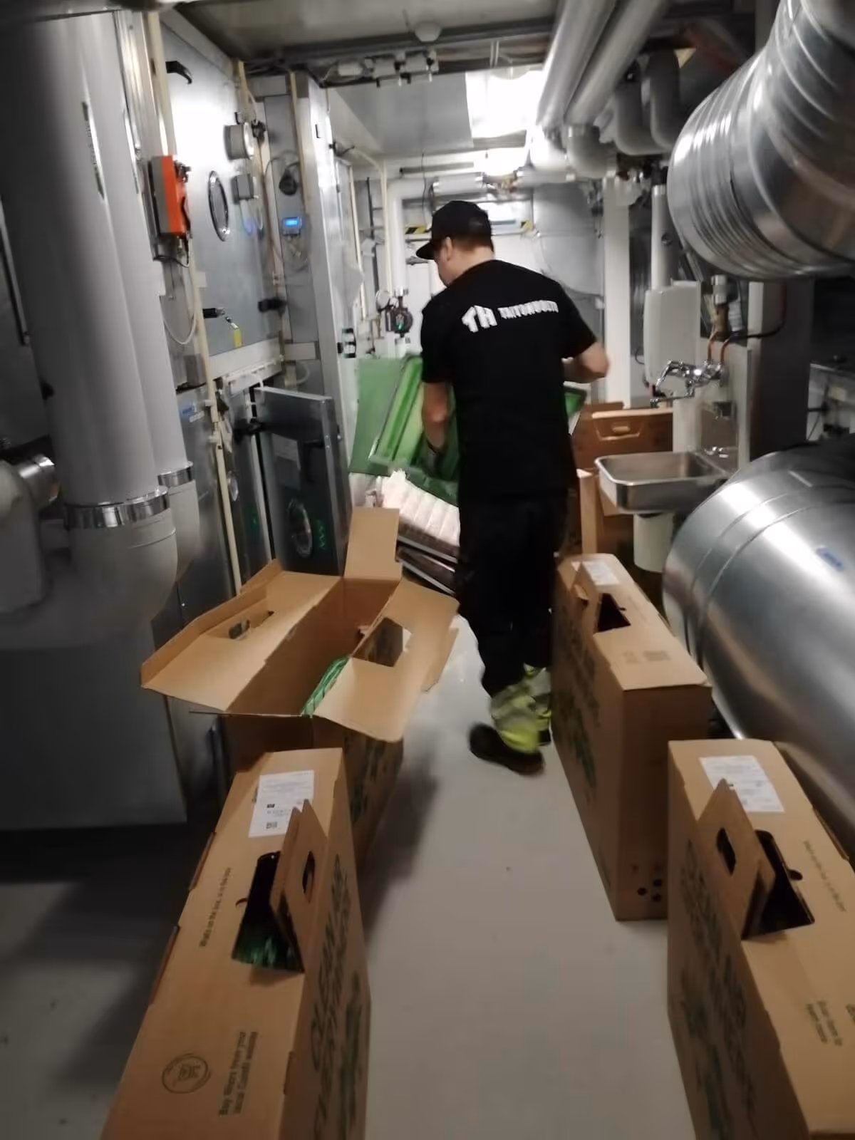 Kuvassa näkyy mies, jolla on musta paita ja lippalakki, kantamassa vihreää esinettä. Hän seisoo huoneessa, jossa on putkia, laatikoita ja teollisuuslaitteita. Valo on kirkas ja tasainen.