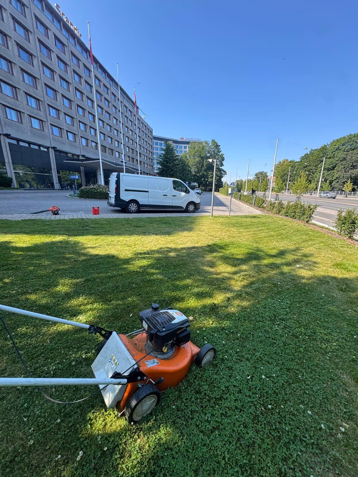 Nurmikkoa leikataan oranssilla ruohonleikkurilla hotellin edessä aurinkoisena päivänä. Taustalla näkyy valkoinen pakettiauto ja rakennuksia.