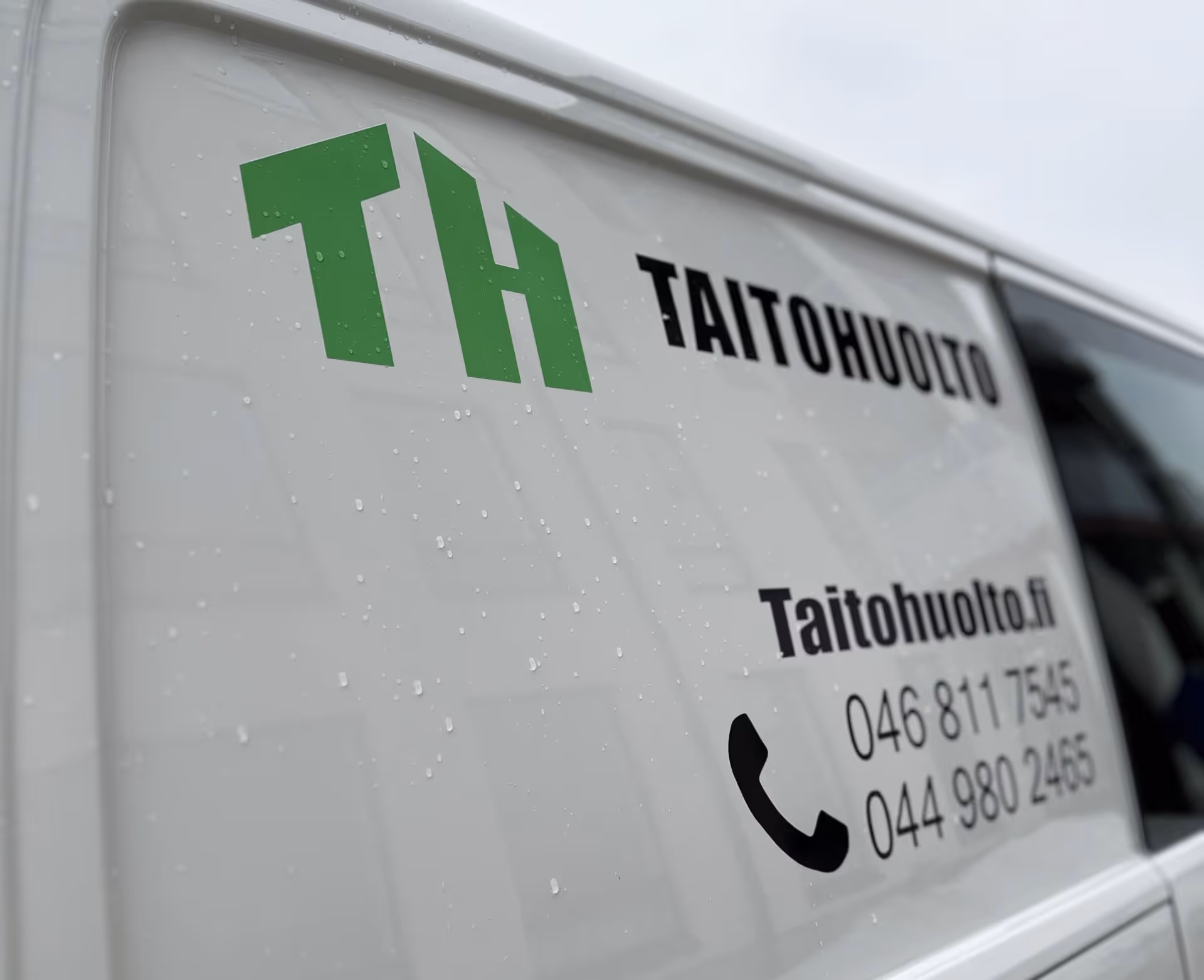 Valkoisen pakettiauton kyljessä on vihreä logo "TH", musta teksti "Taitohuolto" ja "Taitohuolto.fi" sekä puhelinnumerot ja puhelinkuvake.