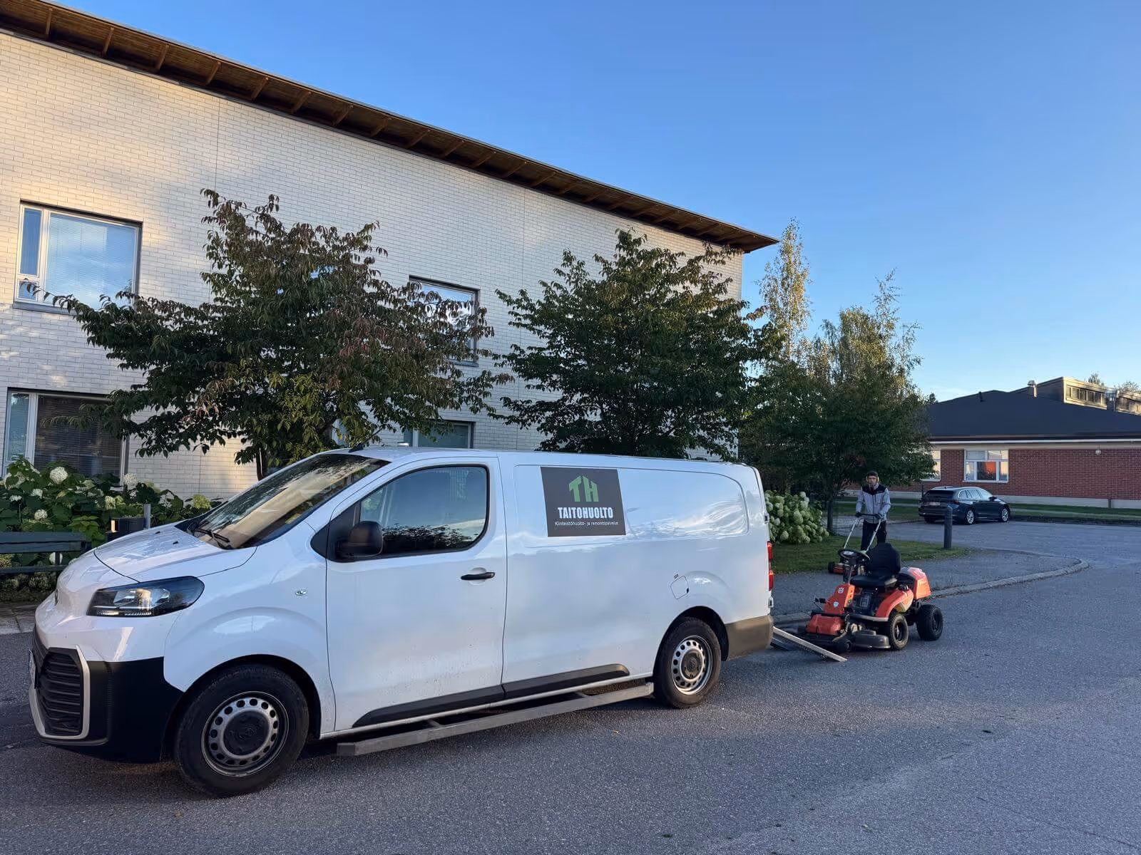 Valkoinen pakettiauto, jonka kyljessä on vihreä logo "TAITOHUOLTO", seisoo kadulla. Punainen ruohonleikkuri on lastaamassa autoon ramppia pitkin. Taustalla on kerrostalo, puita ja henkilö työntämässä ruohonleikkuria. Taivas on sininen.