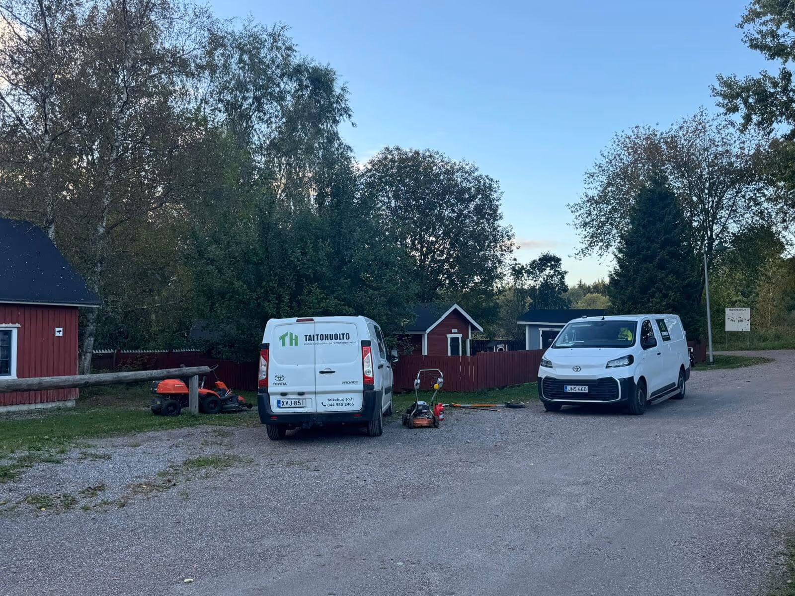 Kuvassa on kaksi valkoista pakettiautoa soraparkkipaikalla, joiden takana on punainen aita ja rakennuksia. Etualalla on ruohonleikkureita. Taivas on sininen ja valaistus on luonnollinen.