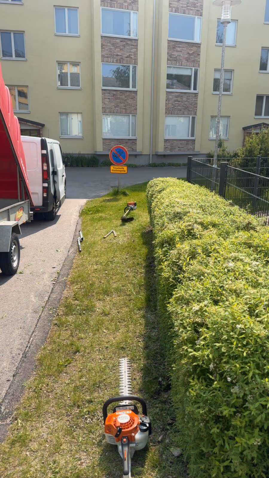 Ulkona on pensasleikkuri, trimmeri ja peräkärry, ja taustalla näkyy rakennus ja liikennemerkki.