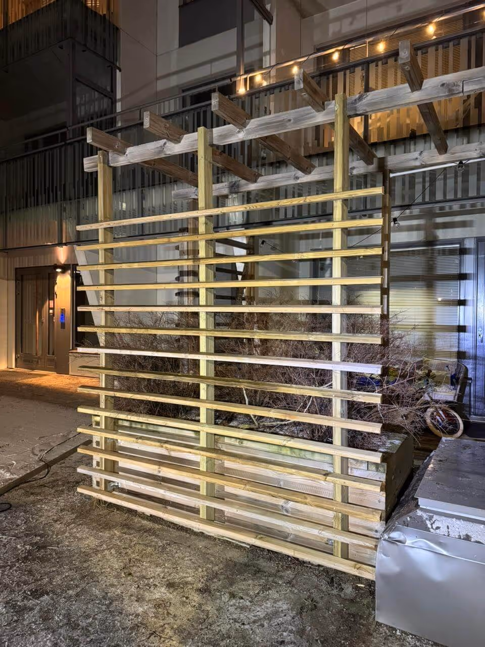 Ulkona on puinen säleikkö, jossa on vaakasuuntaisia lautoja ja pystysuuntaisia tukipylväitä, ja sen yläpuolella on puinen pergola, jossa on valot.