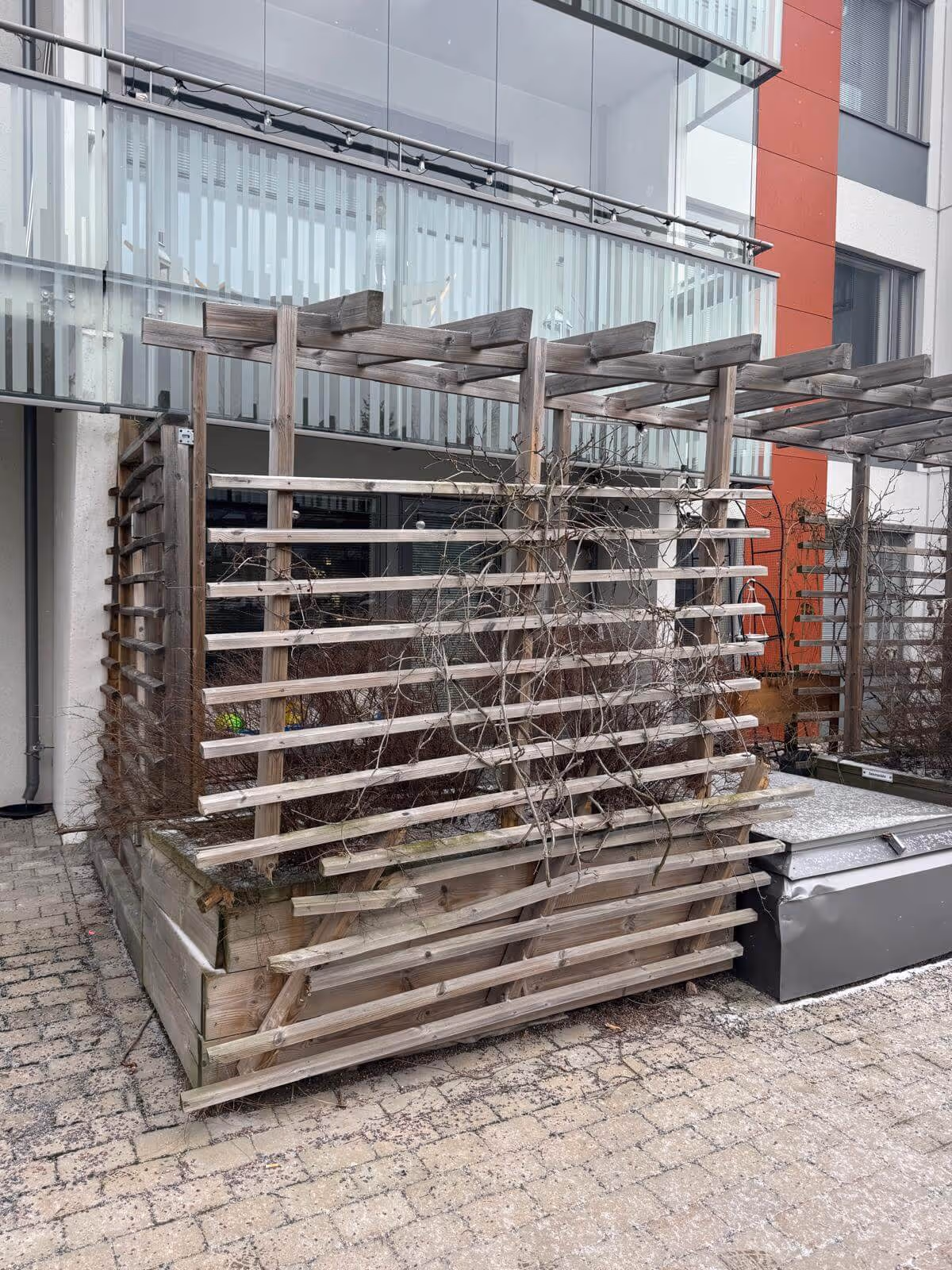 Puinen säleikkölaatikko, jossa on kuivia oksia ja kasveja, sijaitsee rakennuksen ulkopuolella harmaalla säällä.