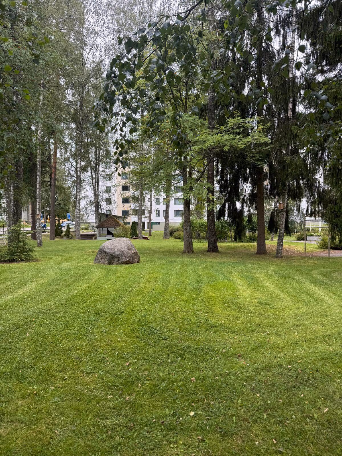 Puistomaisema, jossa on suuri kivi, vihreä nurmikko, puita ja taustalla kerrostalo.