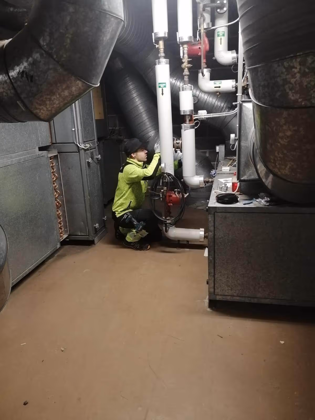 Mies kyykyssä putkien parissa, yllään neonvihreä takki ja musta pipo, huoneessa on metalliputkia ja -kaappeja, valaistus on himmeä.