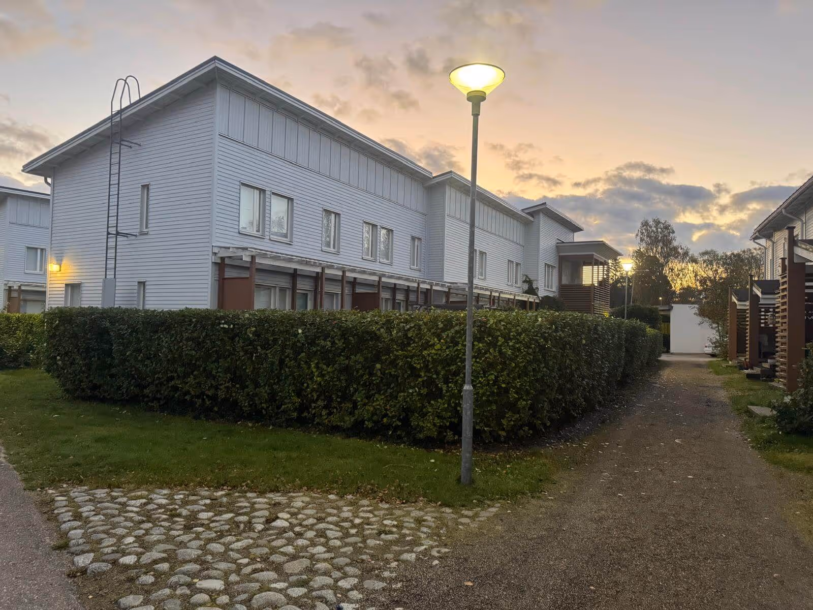 Vaaleanharmaa rakennus, vihreä pensasaita, katuvalaisin ja hämärä taivas.