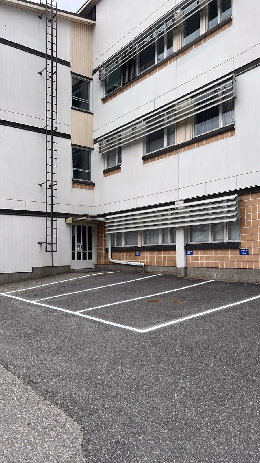 Ulkokuva rakennuksesta, jossa on valkoiset seinät, ikkunat ja metallinen palotikkaat, sekä pysäköintipaikat.