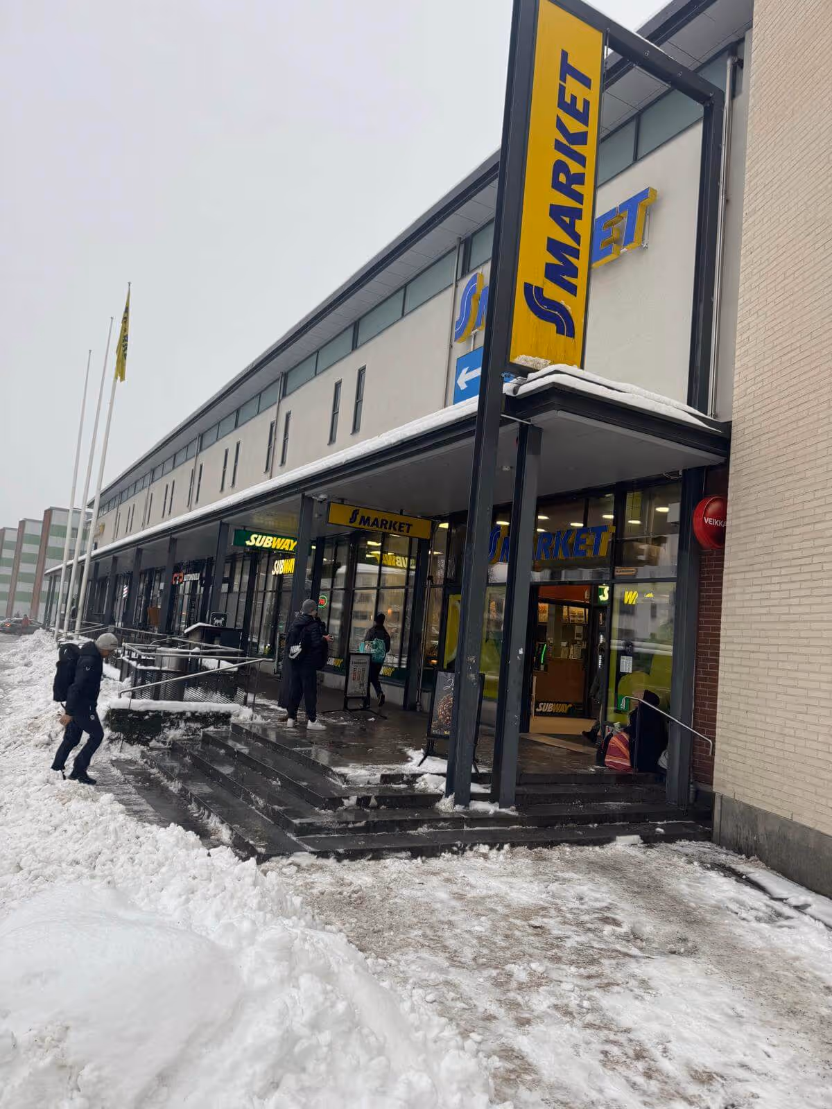 Kuva ulkoa S-marketista talvella, jossa on lunta maassa ja ihmisiä kävelemässä sisään. Rakennuksessa on myös Subway-ravintola. Taivas on harmaa ja valaistus on luonnollinen.