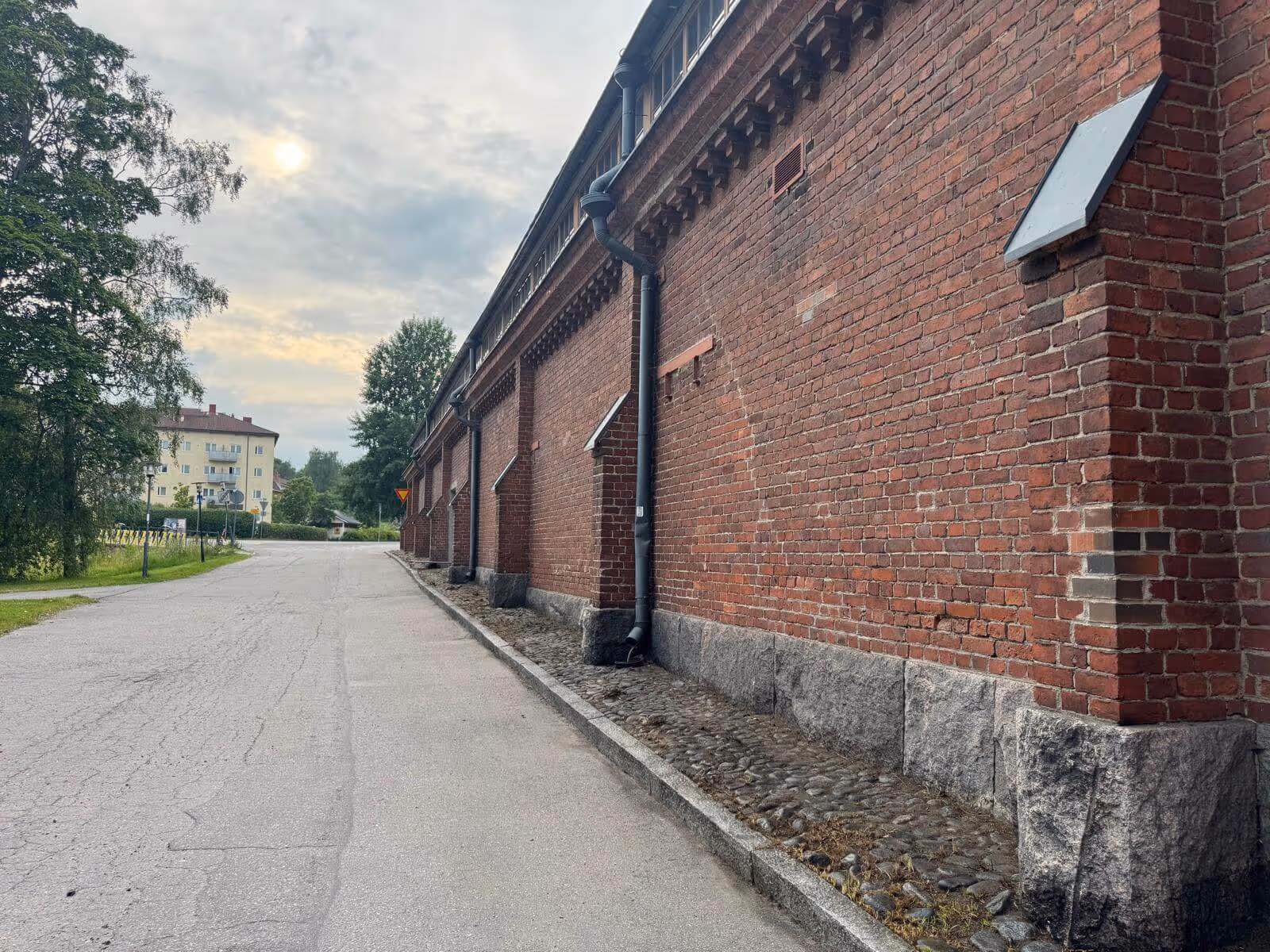 Punatiilinen rakennus kivijalalla reunustaa asfaltoitua katua, taustalla näkyy kerrostalo ja puita, valaistus on pehmeää ja pilvistä.