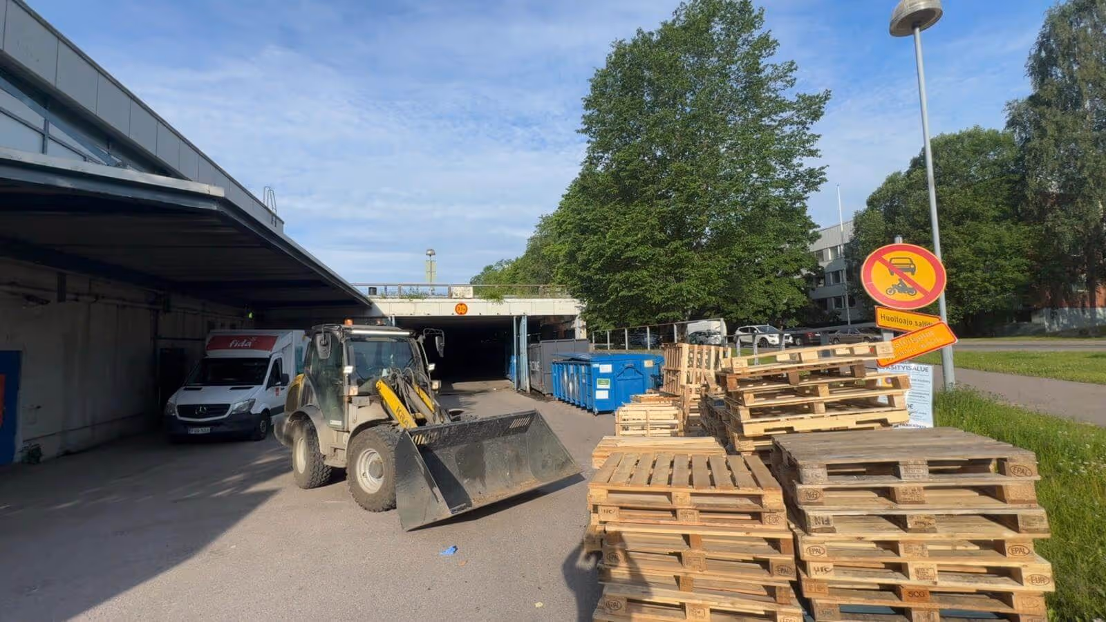 Ulkona on kuva, jossa näkyy traktori, kuormalavoja, pakettiauto ja roskasäiliöitä. Taustalla on vihreitä puita ja sininen taivas. Valo on kirkas ja aurinkoinen.