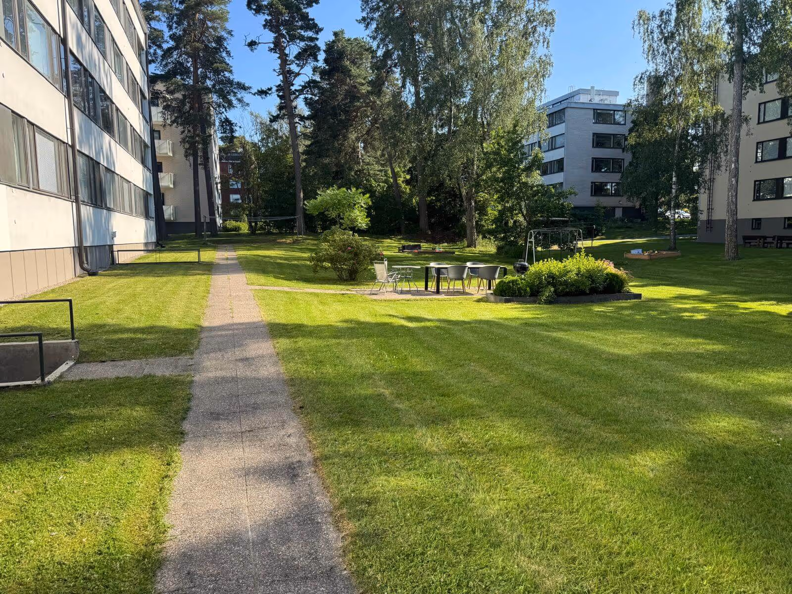 Nurmikkoinen piha-alue, jossa on polku, pöytäryhmä ja leikkivälineitä, valoisana päivänä.